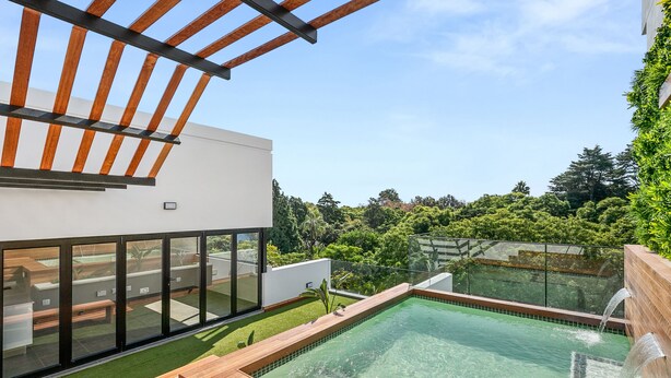 Un área de piscina moderna al aire libre con terraza de madera, barandilla de vidrio y vistas a un jardín exuberante.