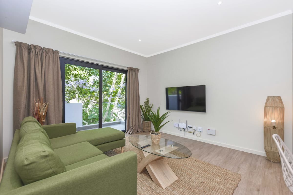 A modern living room with a green sofa, a glass coffee table, a flat-screen TV, and a large window with curtains.