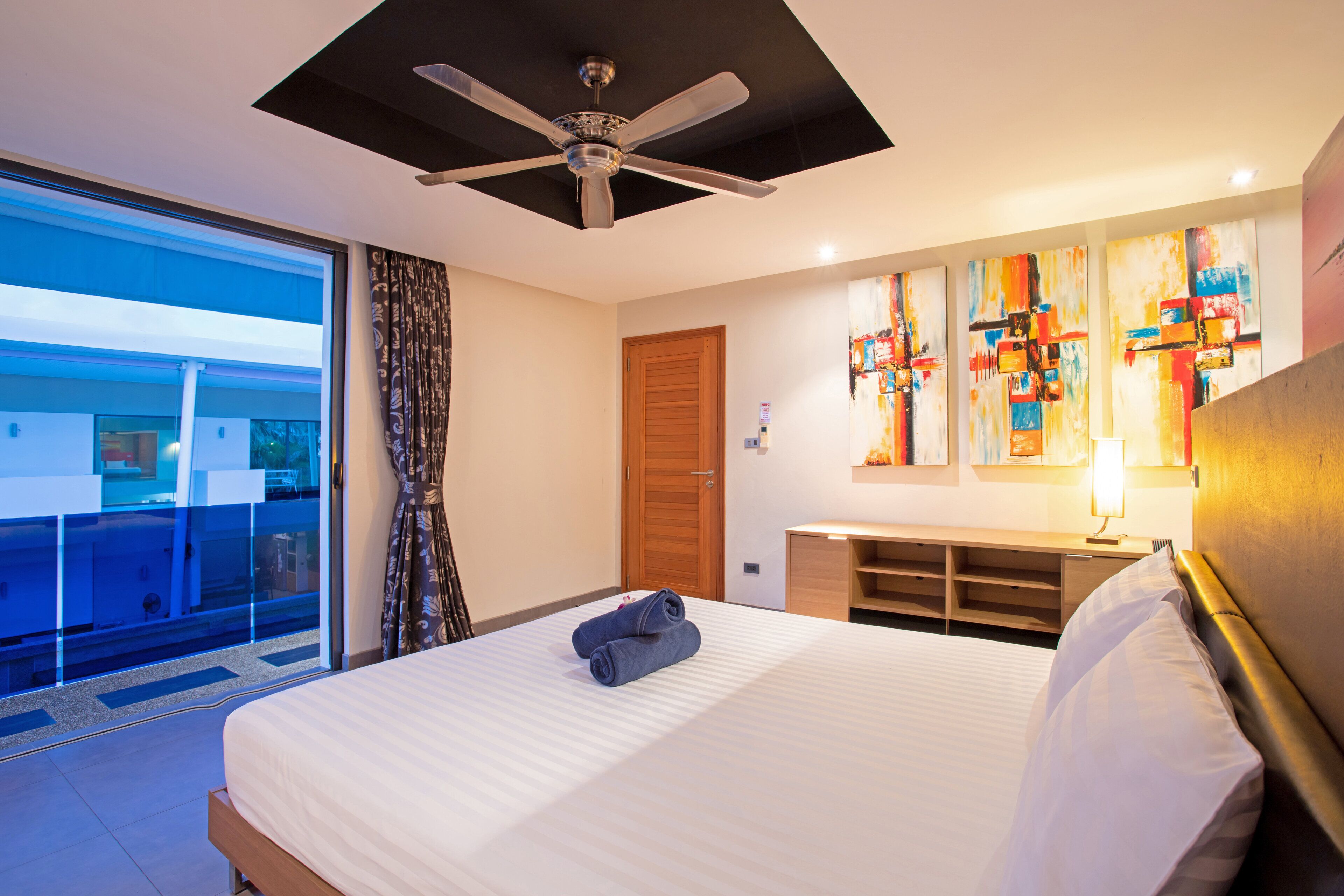 A hotel room with a bed, a ceiling fan, a large window, a door, and artwork on the wall.