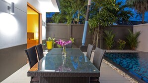 Un espacio moderno para comer al aire libre, con una mesa de vidrio, sillas de mimbre y un jarrón con flores moradas.