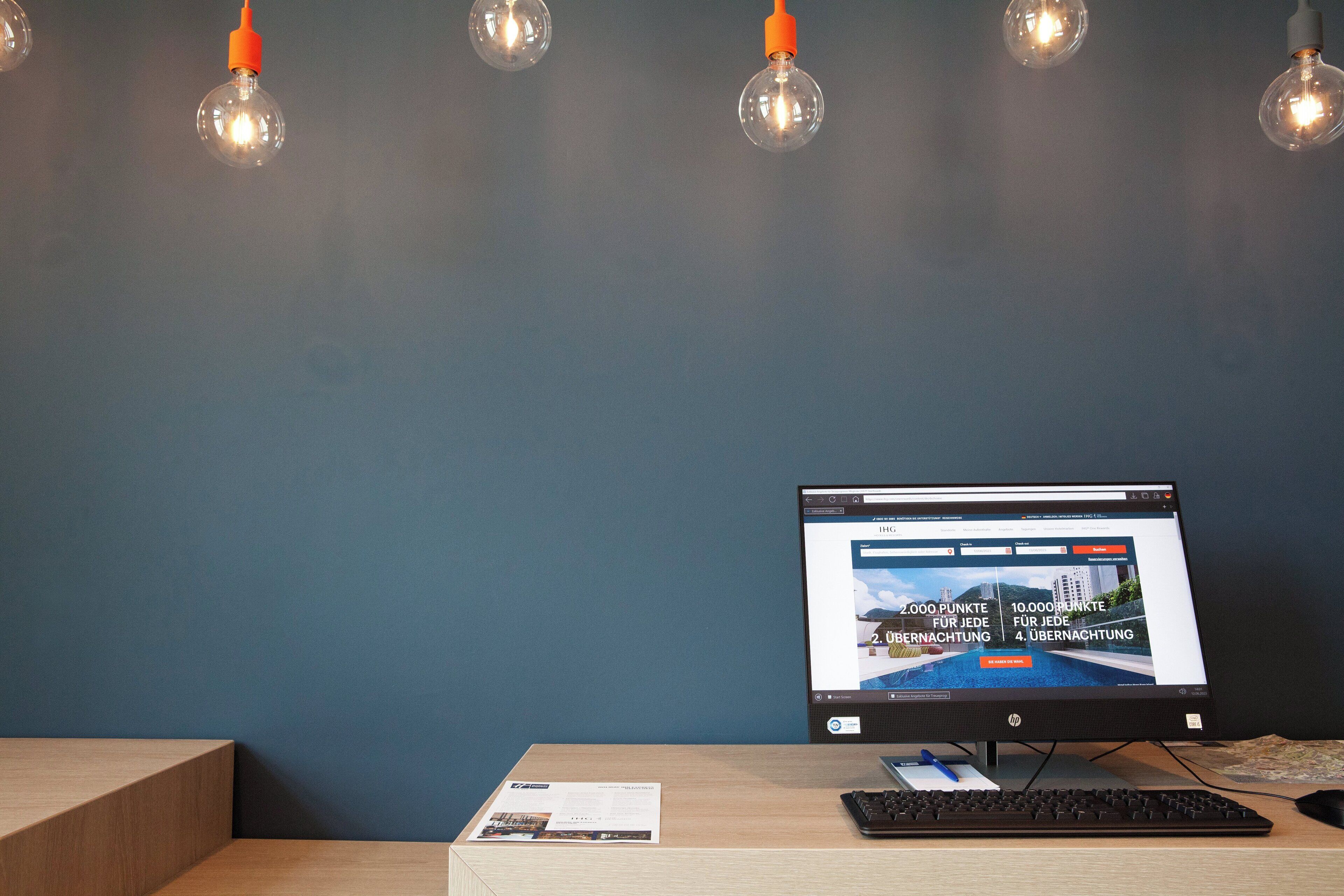 A computer monitor on a desk with a keyboard and a mouse, a brochure, and a wall with hanging light bulbs.