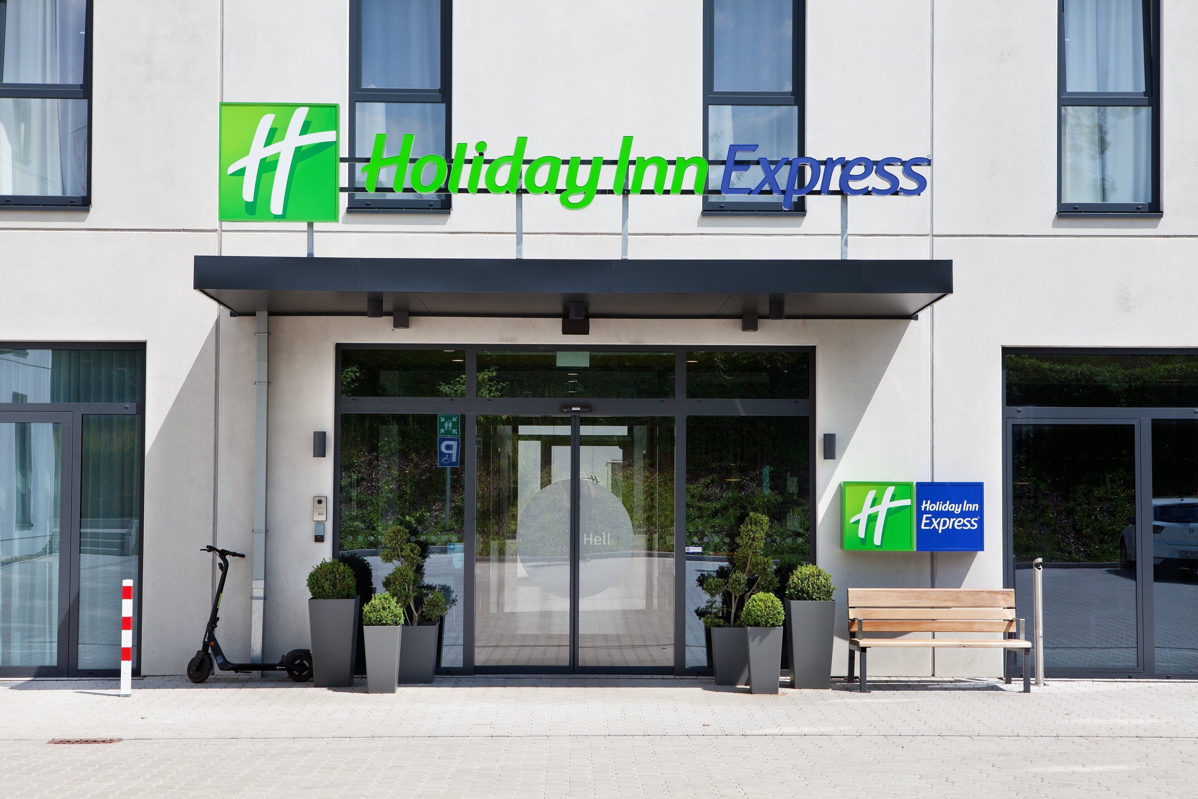 A Holiday Inn Express building with a glass entrance, a scooter parked outside, and a bench in front.