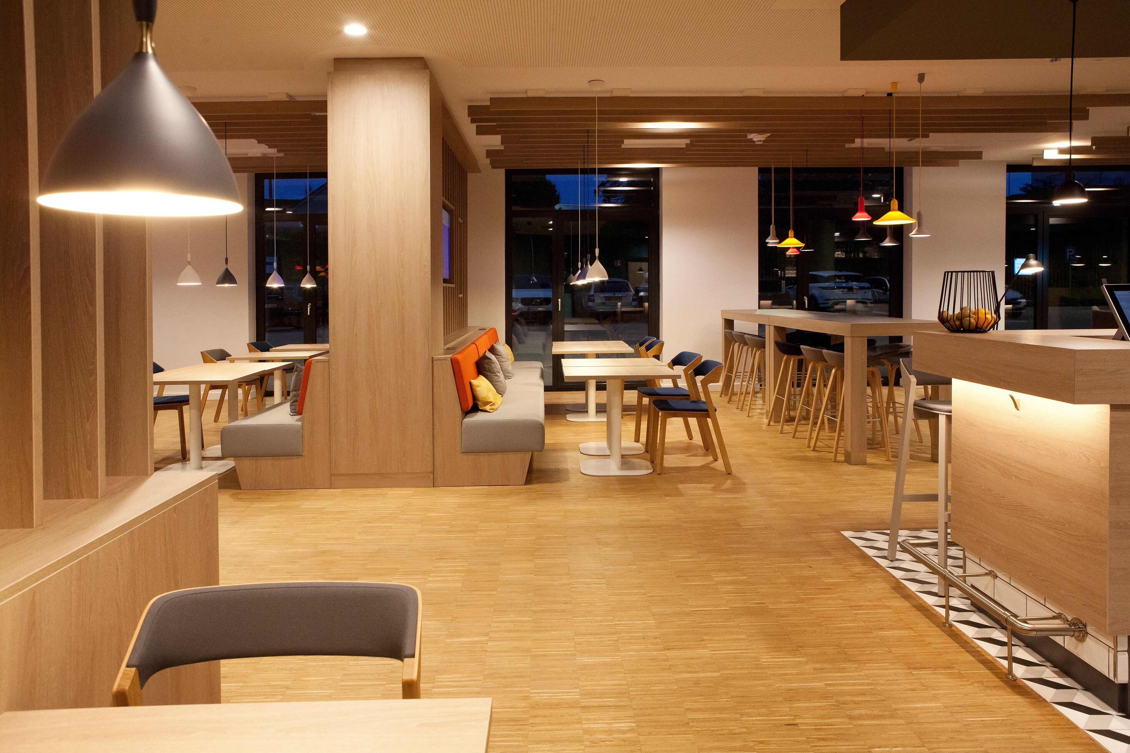 A modern restaurant interior with wooden floors, multiple seating areas, and pendant lighting.