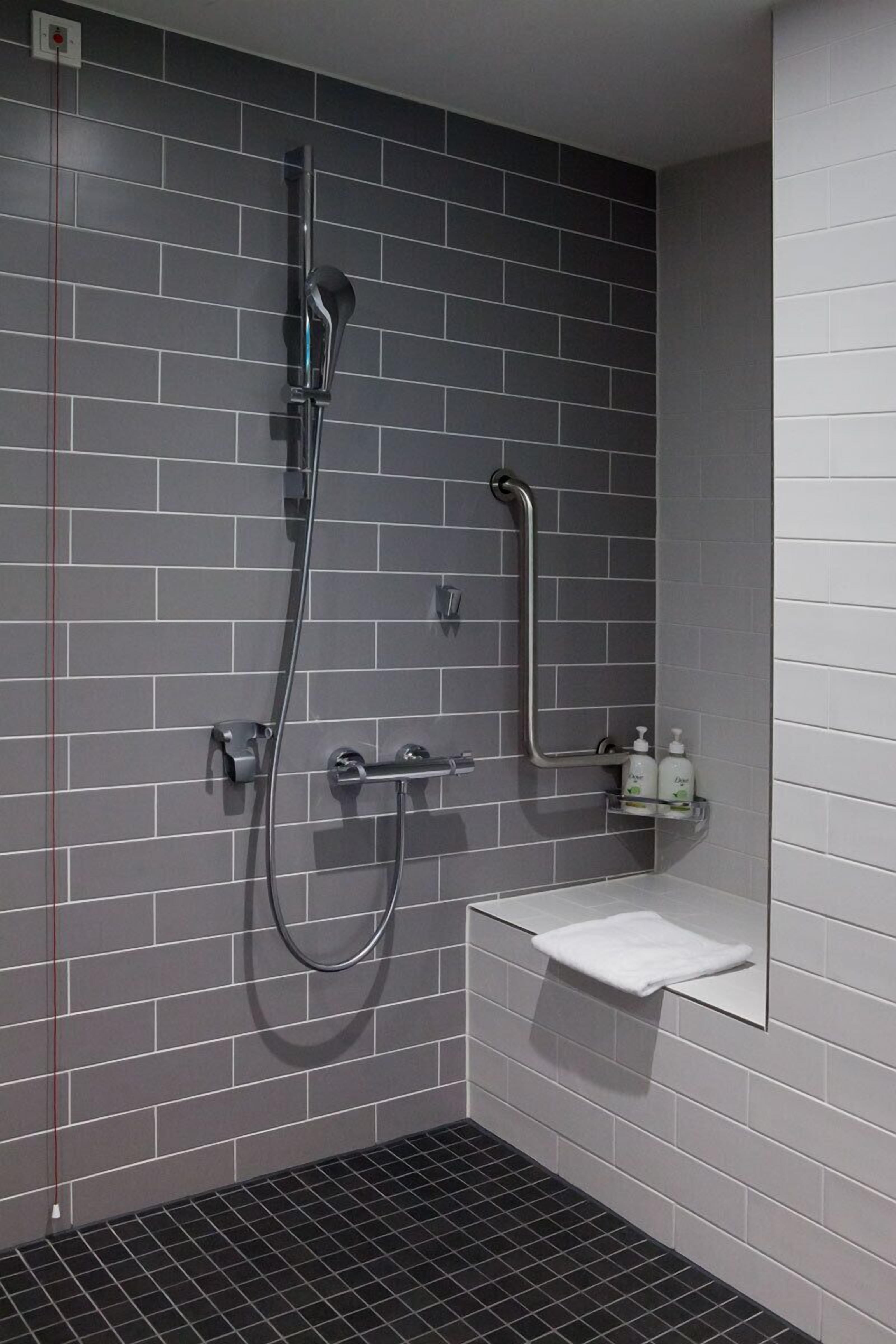 Ein modernes Badezimmer mit grauen Fliesenwänden, einer Duschkabine aus Glas, einem weißen Waschbecken und einer kleinen Ablage mit Pflegeprodukten.