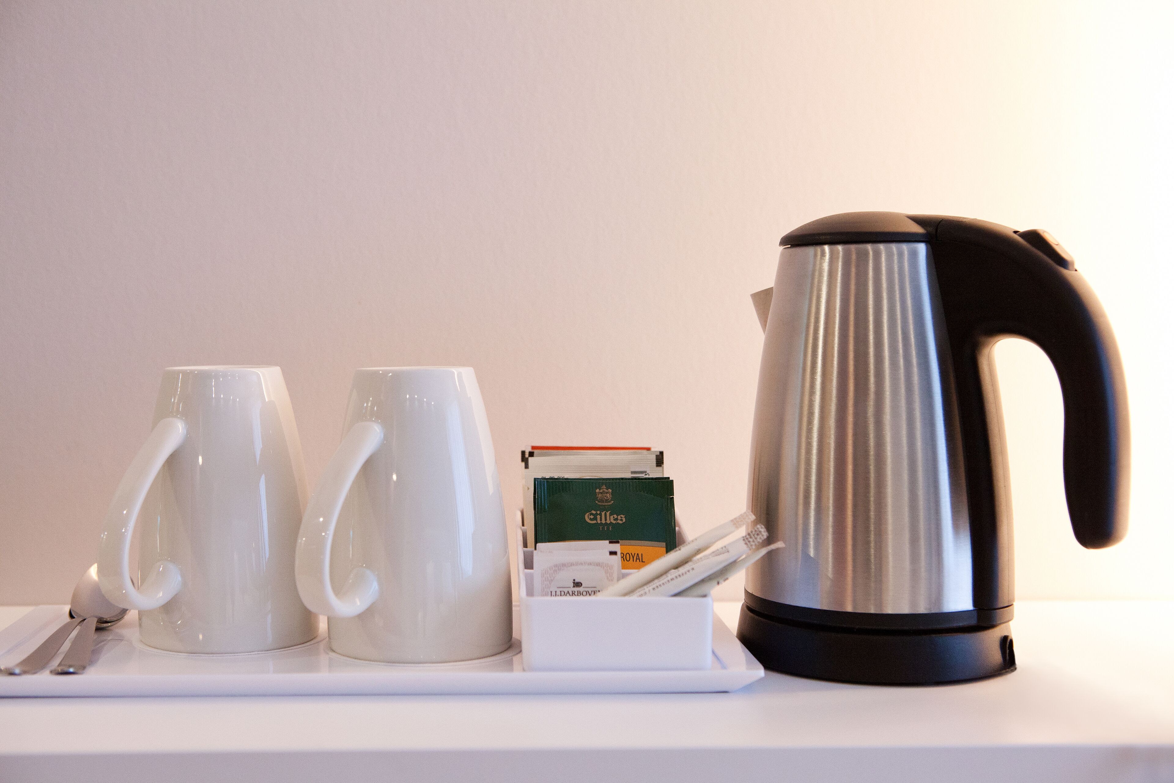 Un plateau blanc avec deux tasses blanches, une bouilloire électrique argentée et une boîte de sachets de thé.