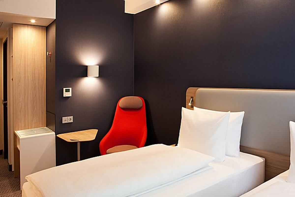 Una habitación de hotel con una cama, una silla roja, una mesita pequeña y una lámpara de pared.