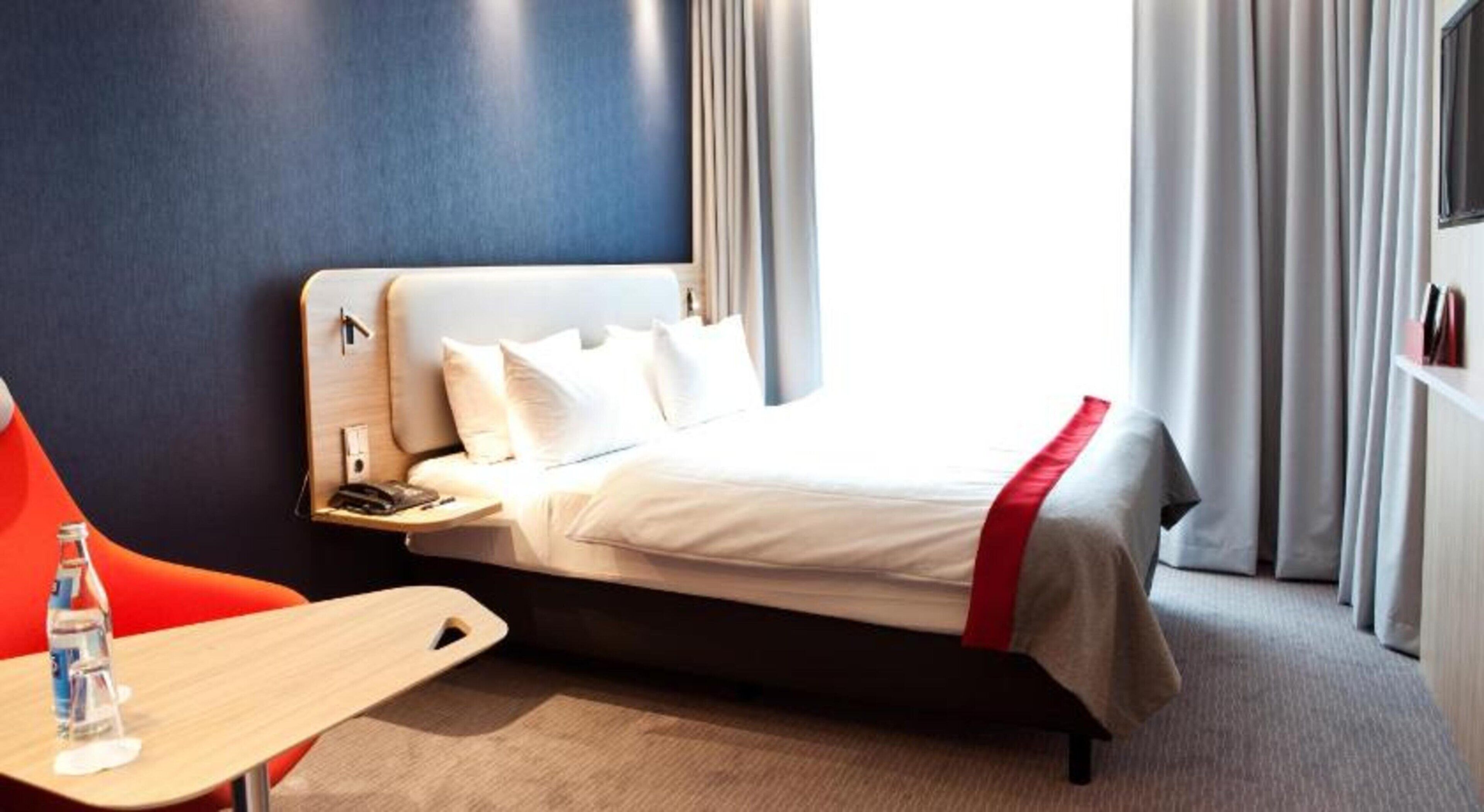 Ein Hotelzimmer mit einem Bett, einem Nachttisch mit Flasche und Gläsern, einem roten Sessel und einem Fenster mit Vorhängen.