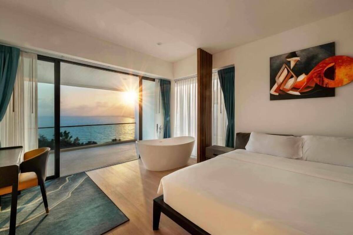 A modern hotel room with a large bed, a bathtub, a dining area, and a view of the ocean.
