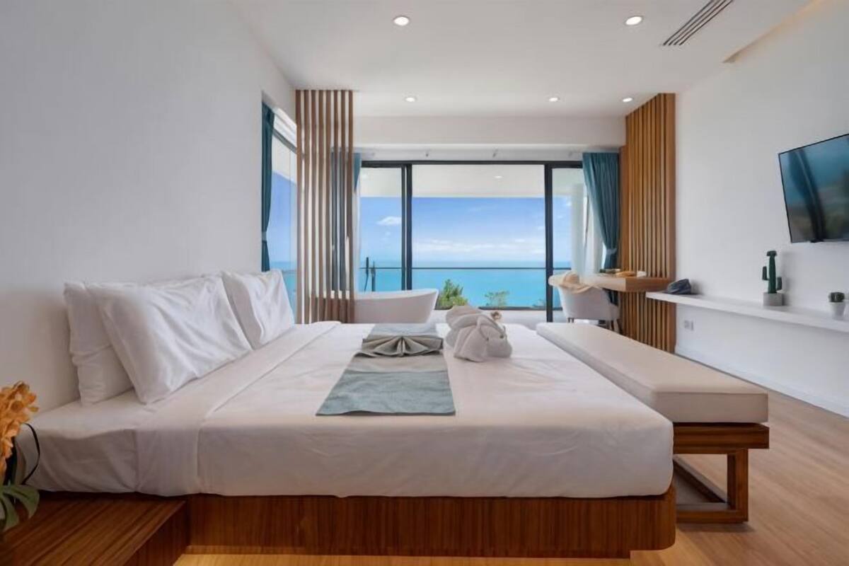 A modern bedroom with a large bed, a view of the sea, a bathtub, and a TV on the wall.