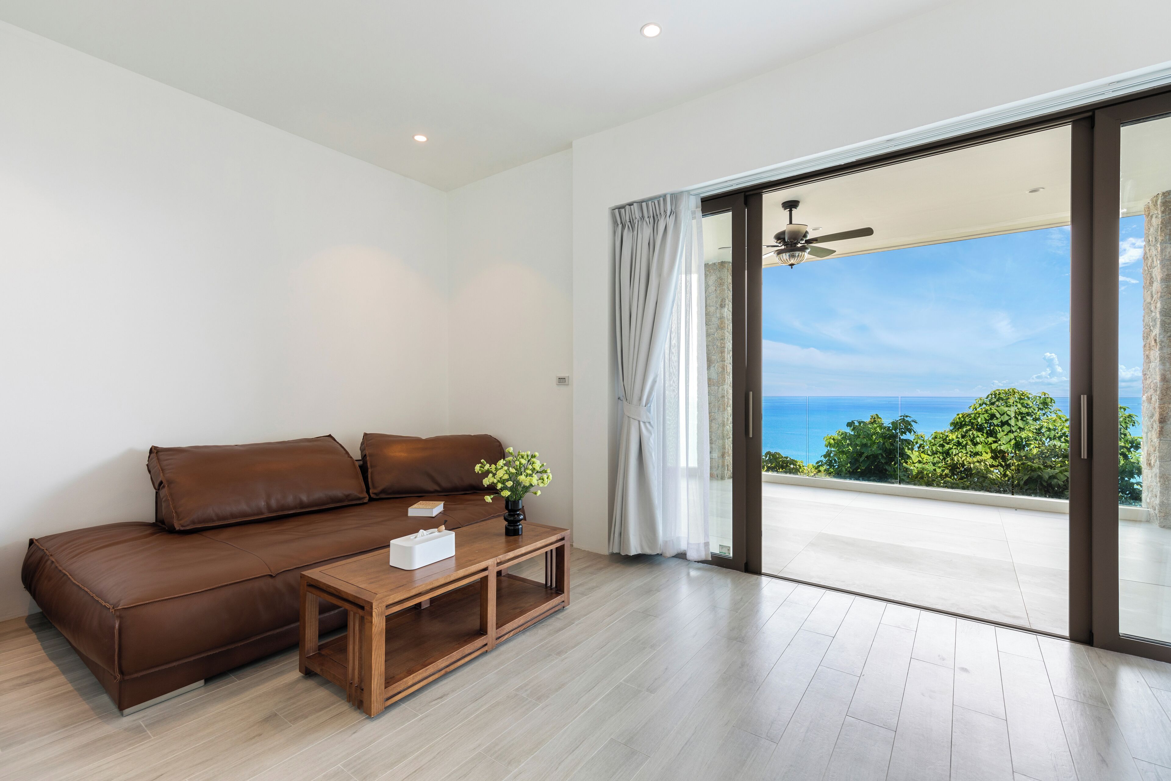 A modern living room with a brown leather sofa, a wooden coffee table, and a view of the ocean through large glass doors.