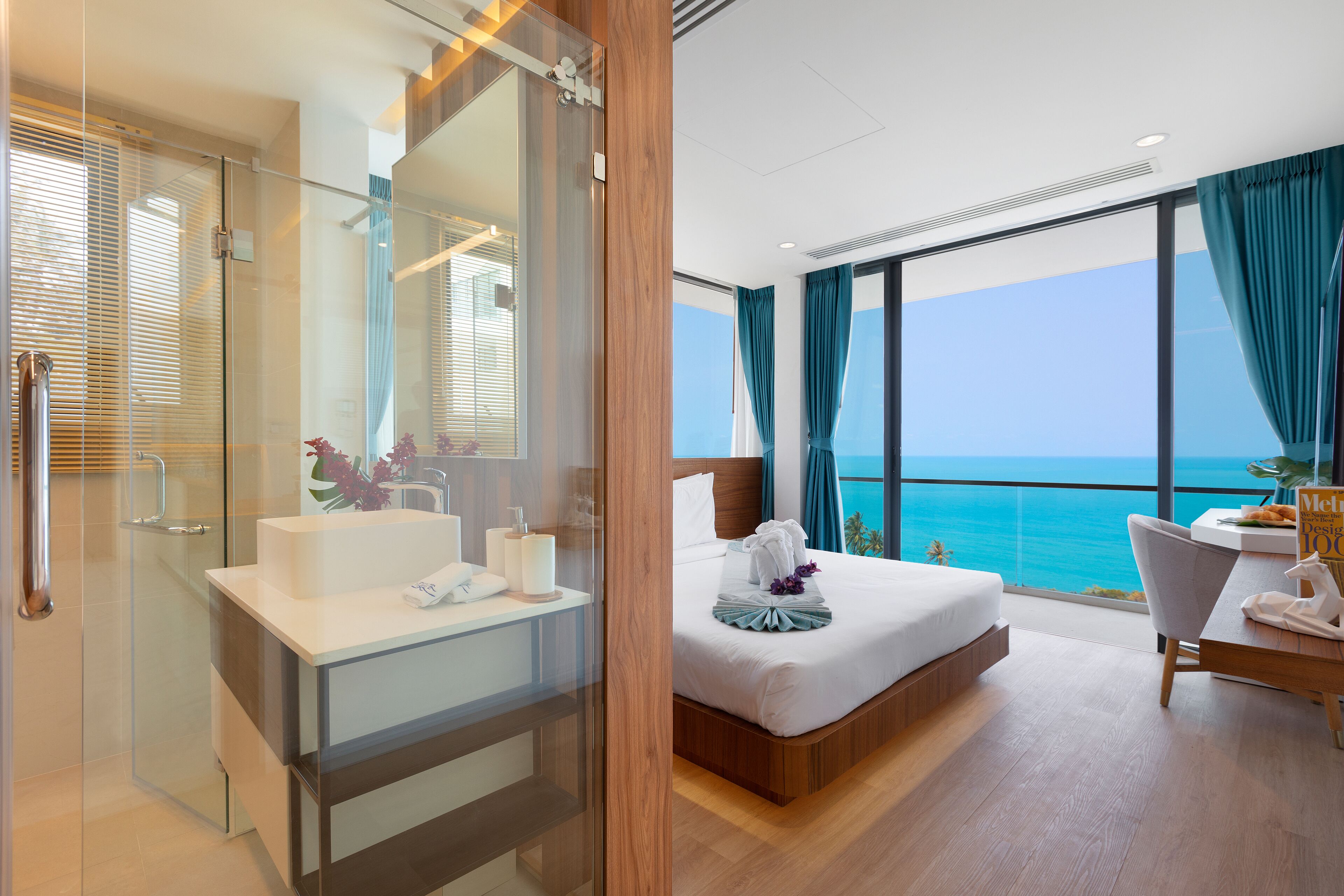 A modern hotel room with a glass shower enclosure, a large bed with a view of the ocean, and a wooden desk with a chair.