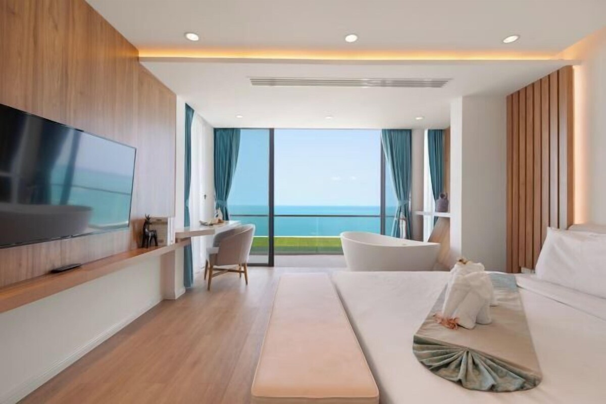 A modern hotel room with a large TV, a desk, a chair, a bathtub, and a bed.