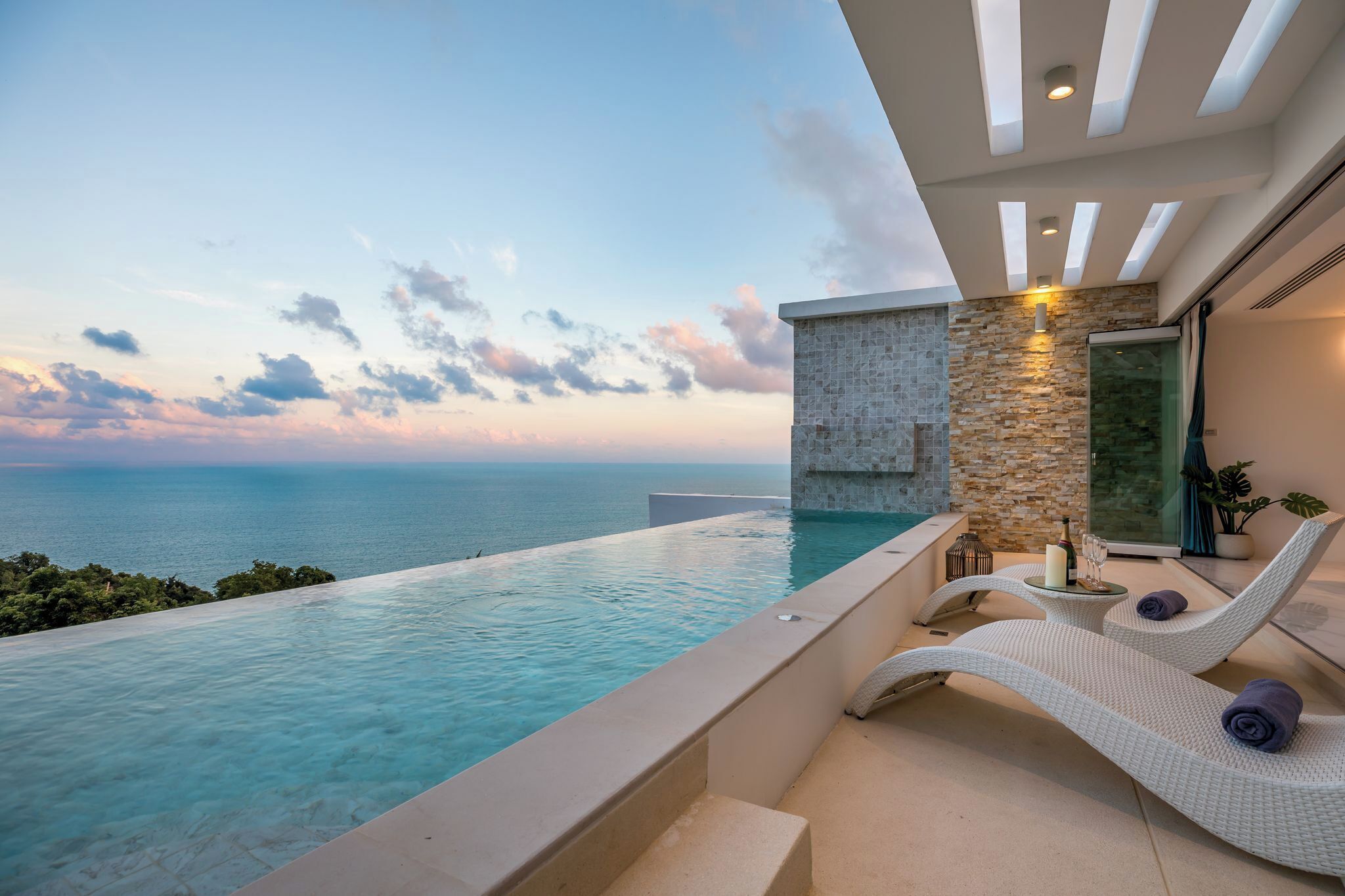 En modern infinitypool med fri utsikt över havet, en stenmur och en glasdörr som leder ut till en balkong med solstolar och ett litet bord med drycker.