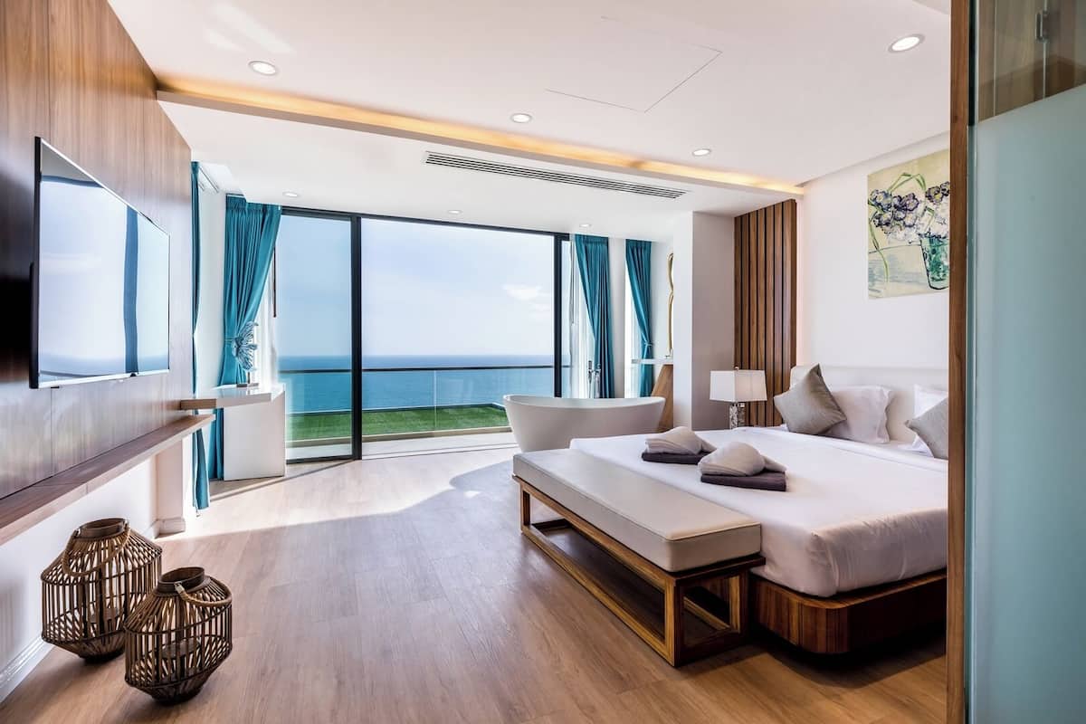A modern hotel room with a large bed, a seating area, and a balcony with a view of the sea.
