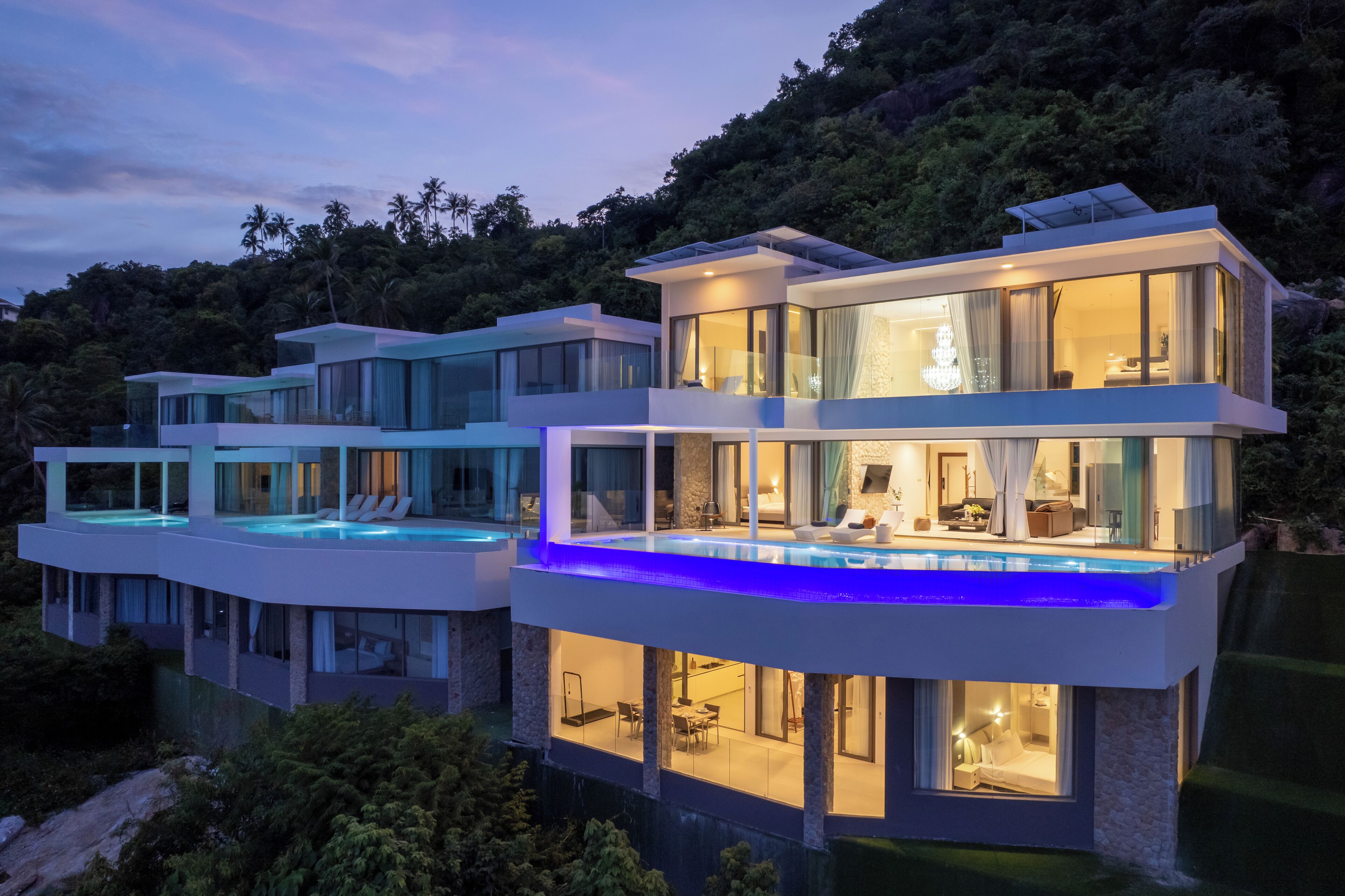 A modern multi-story house with a swimming pool on the upper level, surrounded by lush greenery.
