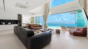 A modern living room with a large black leather sofa, a brown leather couch, and a glass dining table. There is a view of the ocean through large windows. - Villa The Wave 2 Residence (Koh Samui)