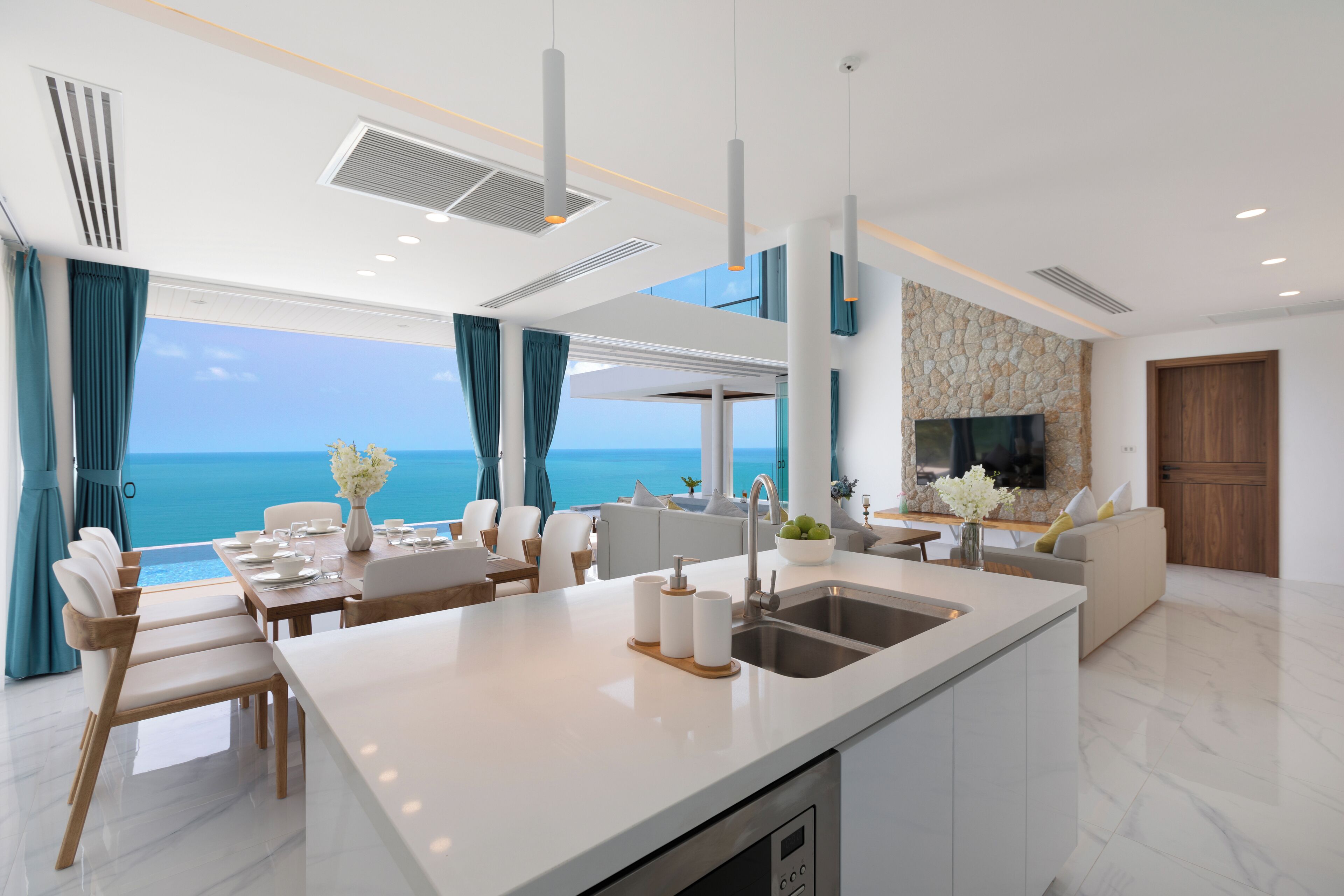 A modern kitchen with a central island, a dining area with a view of the sea, and a living area with a stone accent wall and a TV.