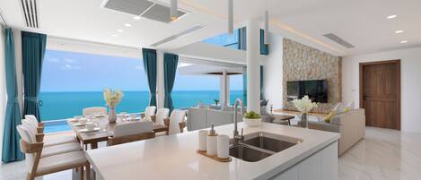 A modern kitchen with a central island, a dining area with a view of the sea, and a living area with a stone accent wall and a TV.