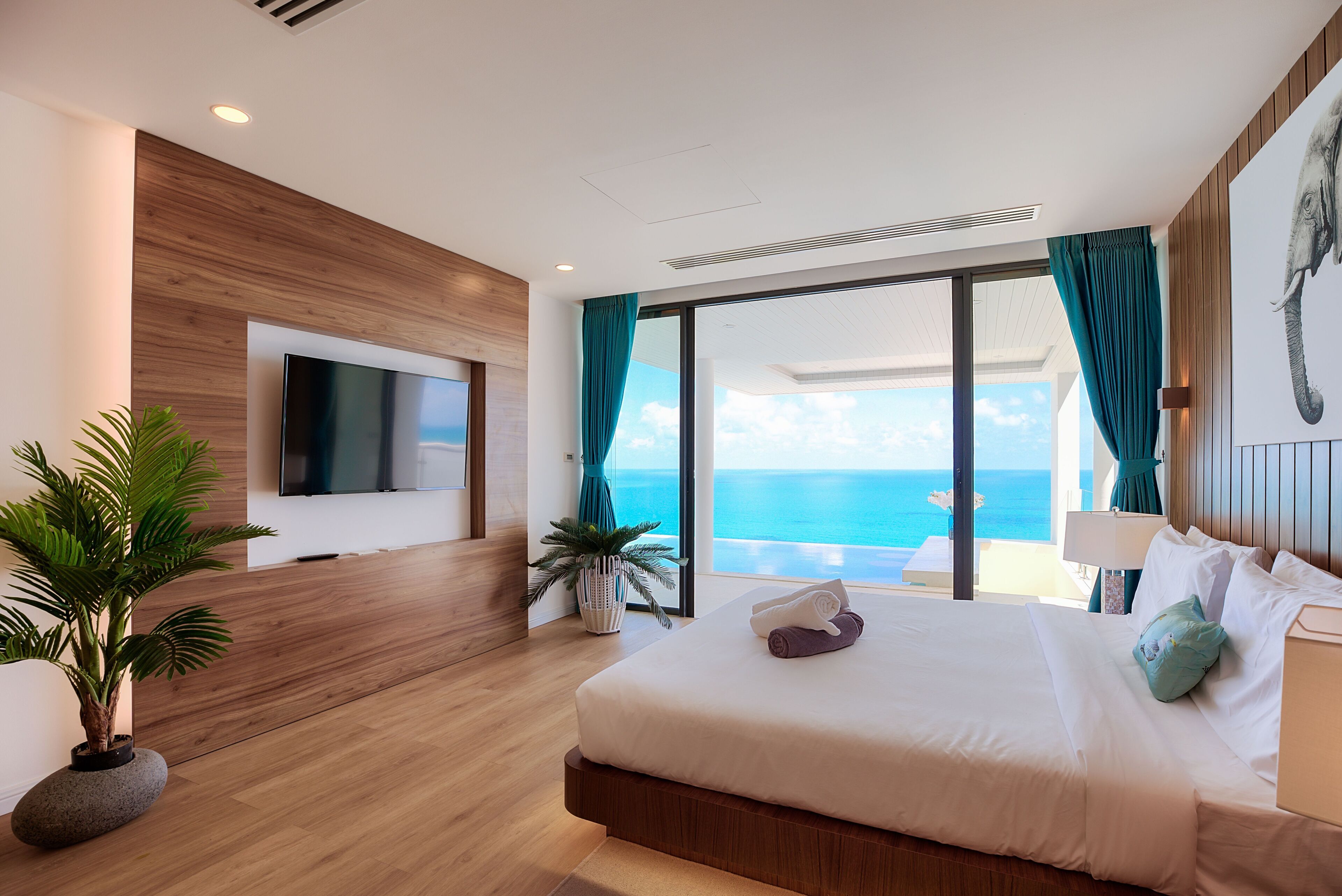 A modern hotel room with a large bed, a flat-screen TV, a potted plant, and a view of the ocean.