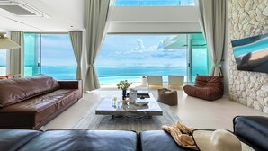 A modern living room with a large window overlooking the sea, a leather sofa, a coffee table, and a stone accent wall. - Villa The Wave 2 Residence (Koh Samui)