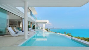 A modern infinity pool with a clear view of the ocean, a covered outdoor area with lounge chairs, and a large white building with glass walls. - Villa The Wave 2 Residence (Koh Samui)