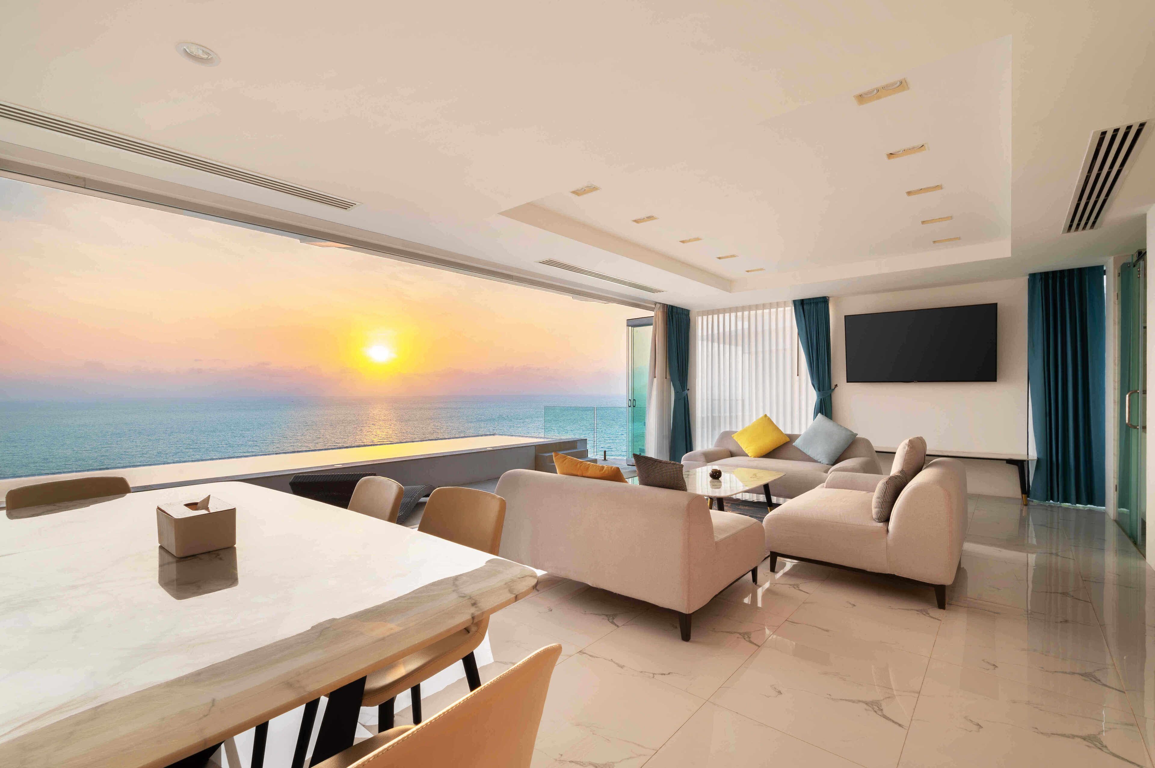 Ein modernes Hotelzimmer mit großem Fenster, das einen Sonnenuntergang über dem Meer zeigt, einem Flachbildfernseher, einer Couch, einer Essecke und einem sauberen, gefliesten Boden.