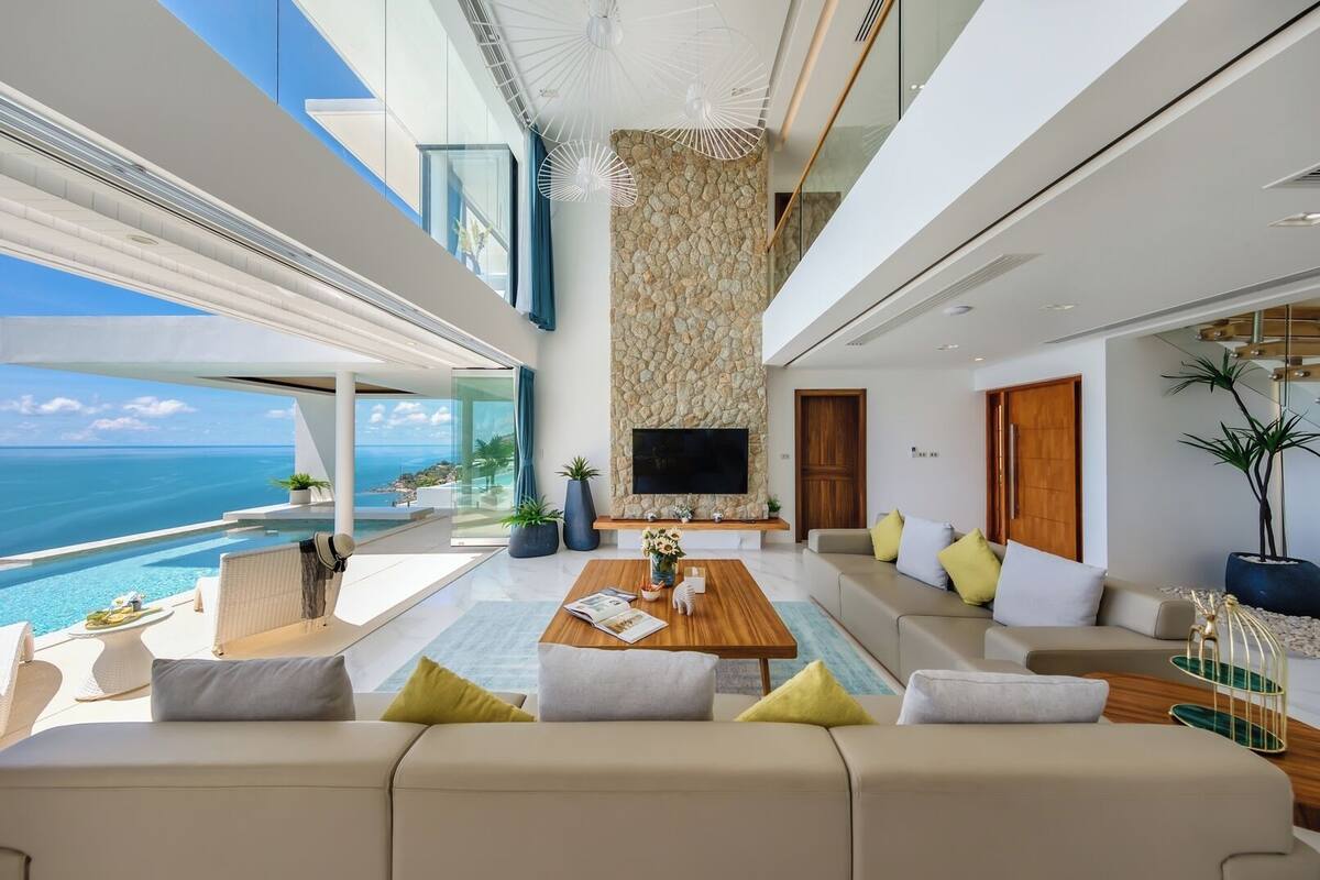 A modern living room with a large sofa, a coffee table, and a TV mounted on a stone wall. There is a view of the ocean through large windows.