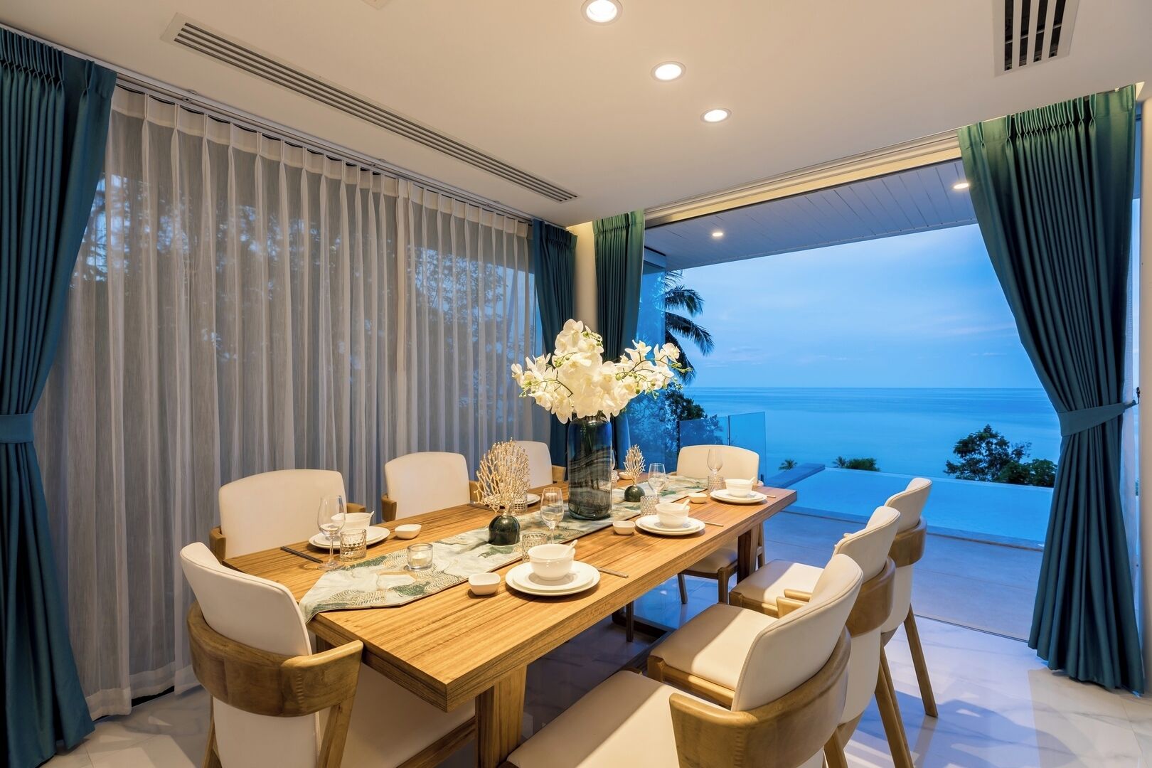 Uma sala de jantar com uma mesa de madeira posta para seis pessoas, rodeada por cadeiras brancas. Há um vaso com flores brancas sobre a mesa, e o quarto possui grandes janelas com cortinas.