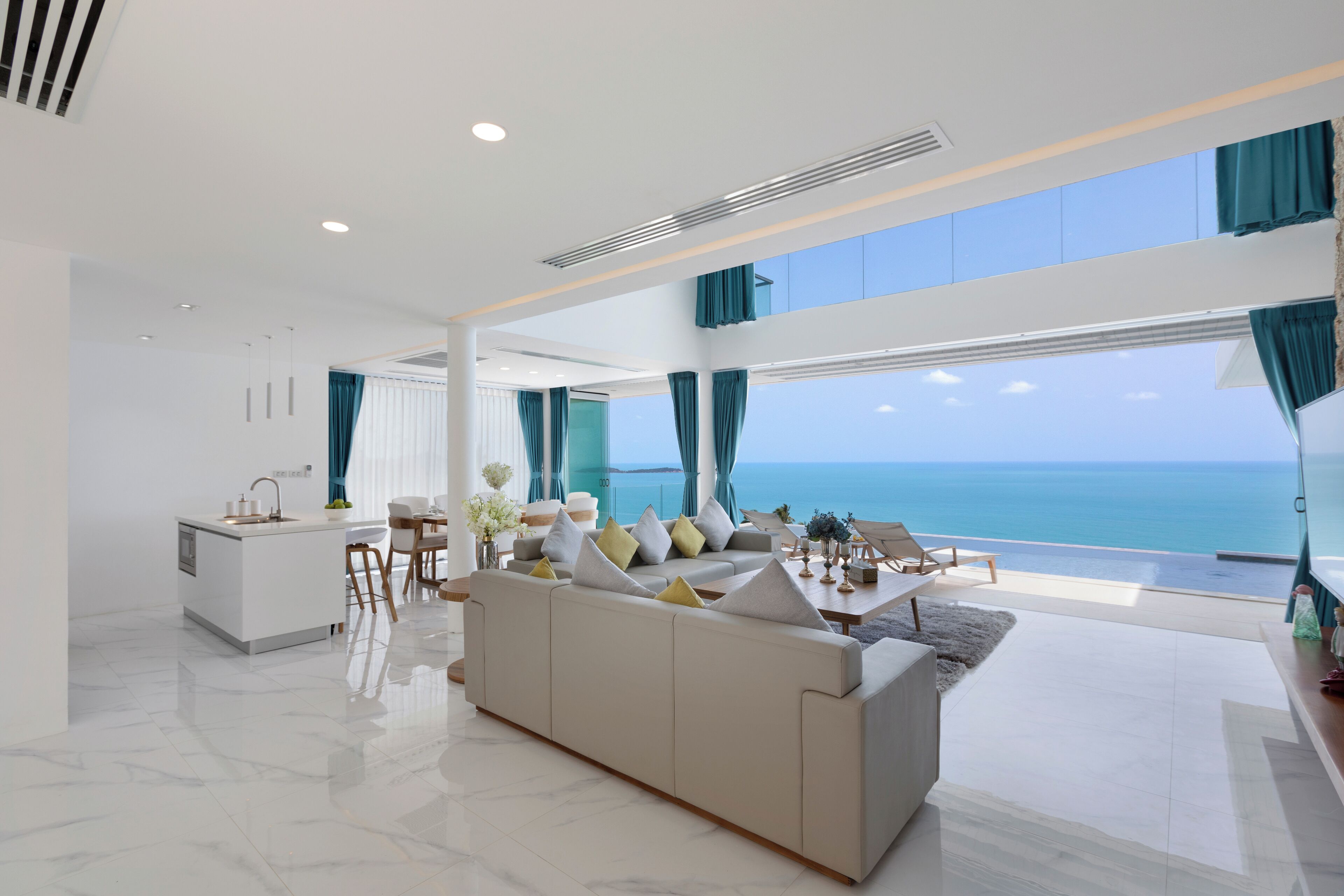 A modern living room with a large sofa, a dining area, and a kitchen. The room has large windows offering a view of the sea.