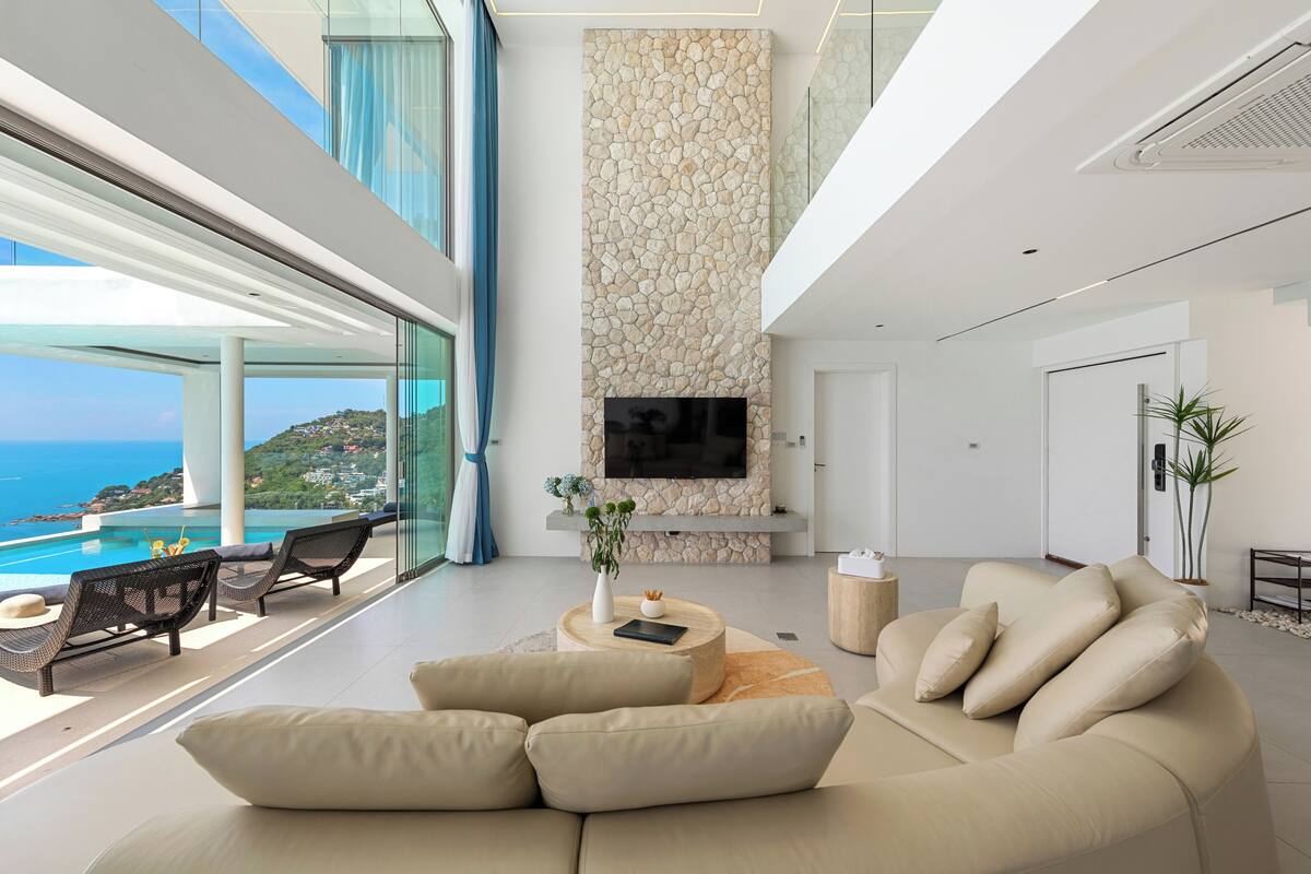 A modern living room with a large sofa, a flat-screen TV mounted on a stone wall, and a view of the ocean.