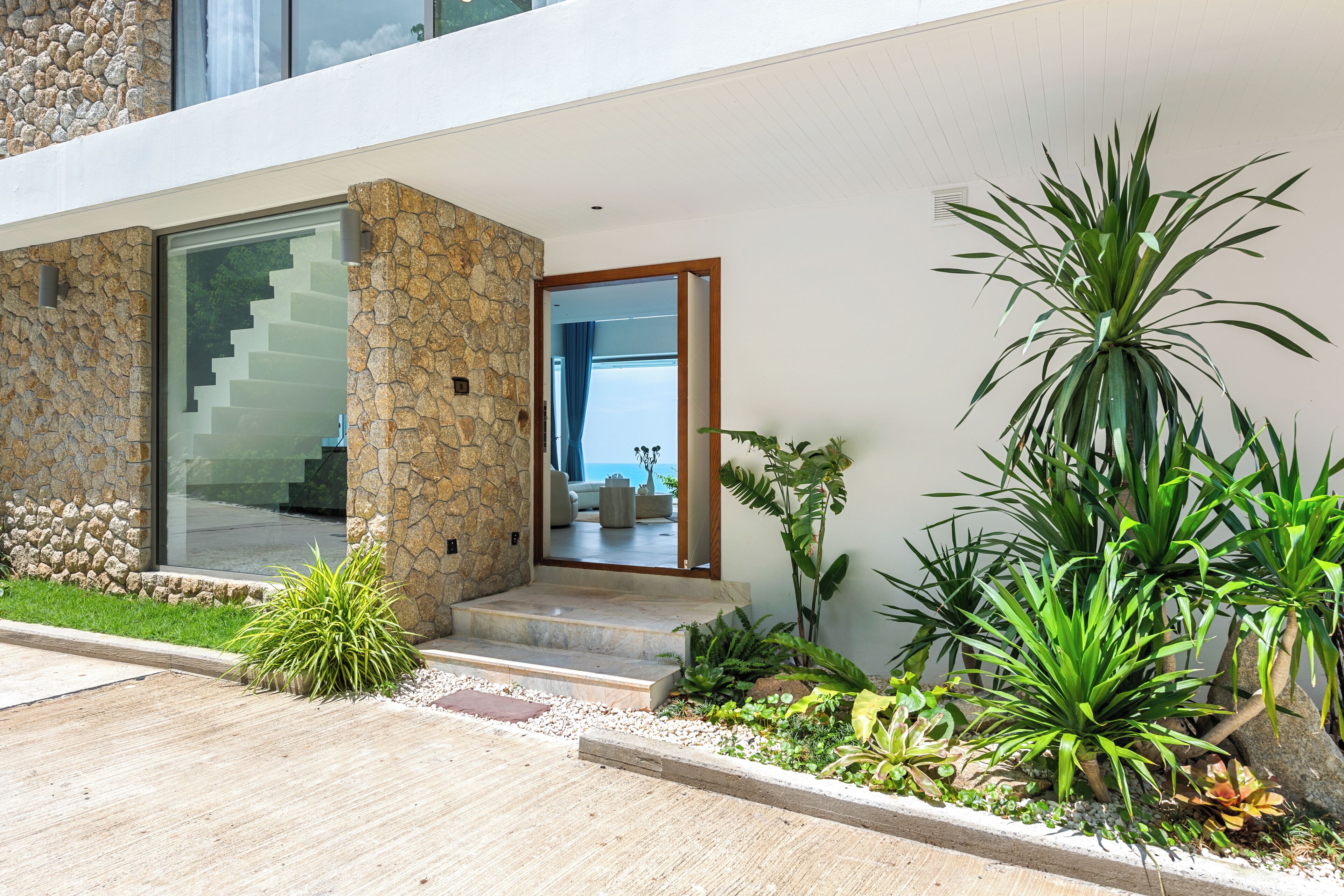 Une maison moderne avec une façade en pierre, une porte vitrée donnant sur un balcon et une variété de plantes.