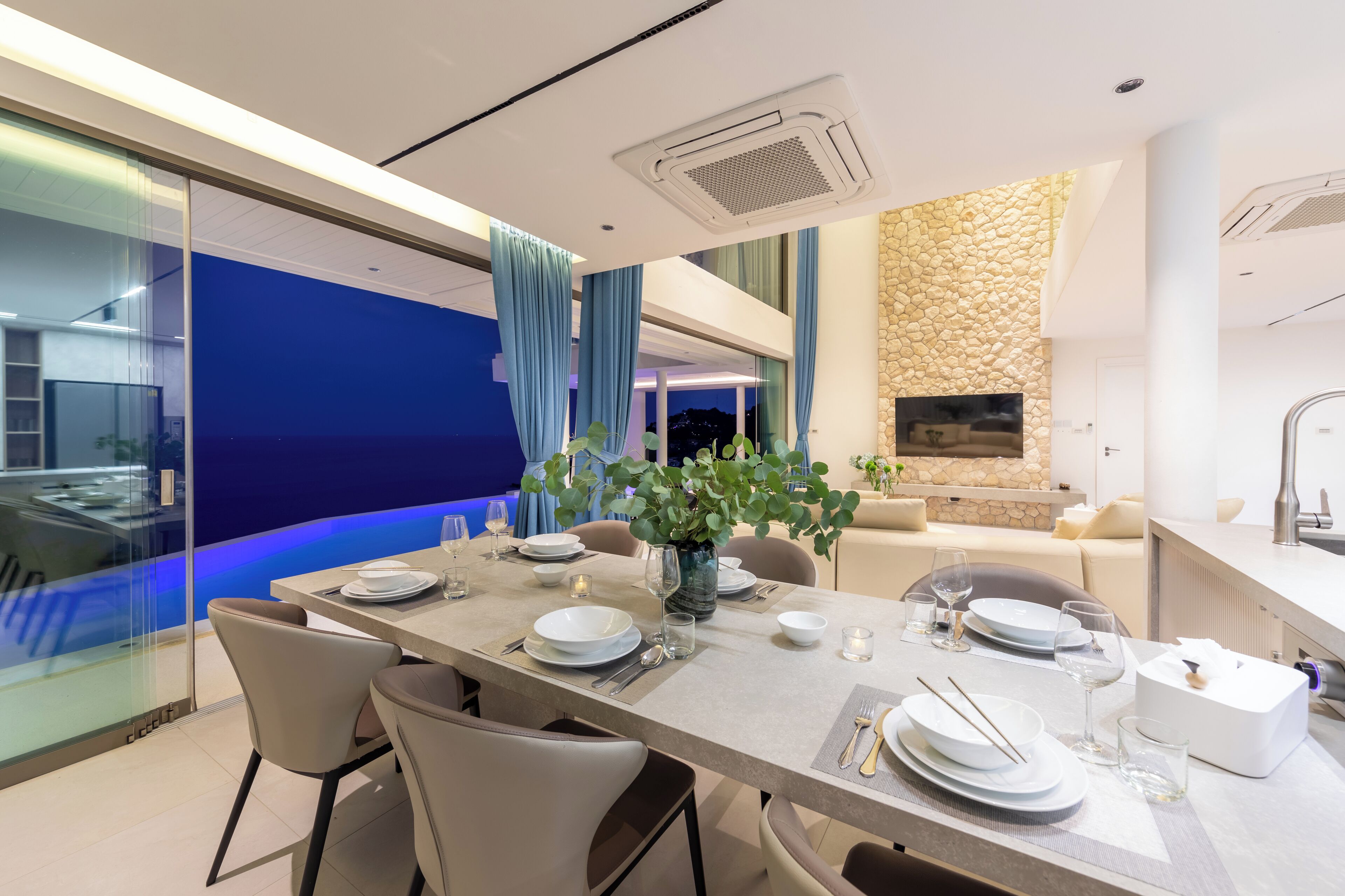 A modern dining area with a long table set for four, glass walls, a stone accent wall, and a view of the city at night.