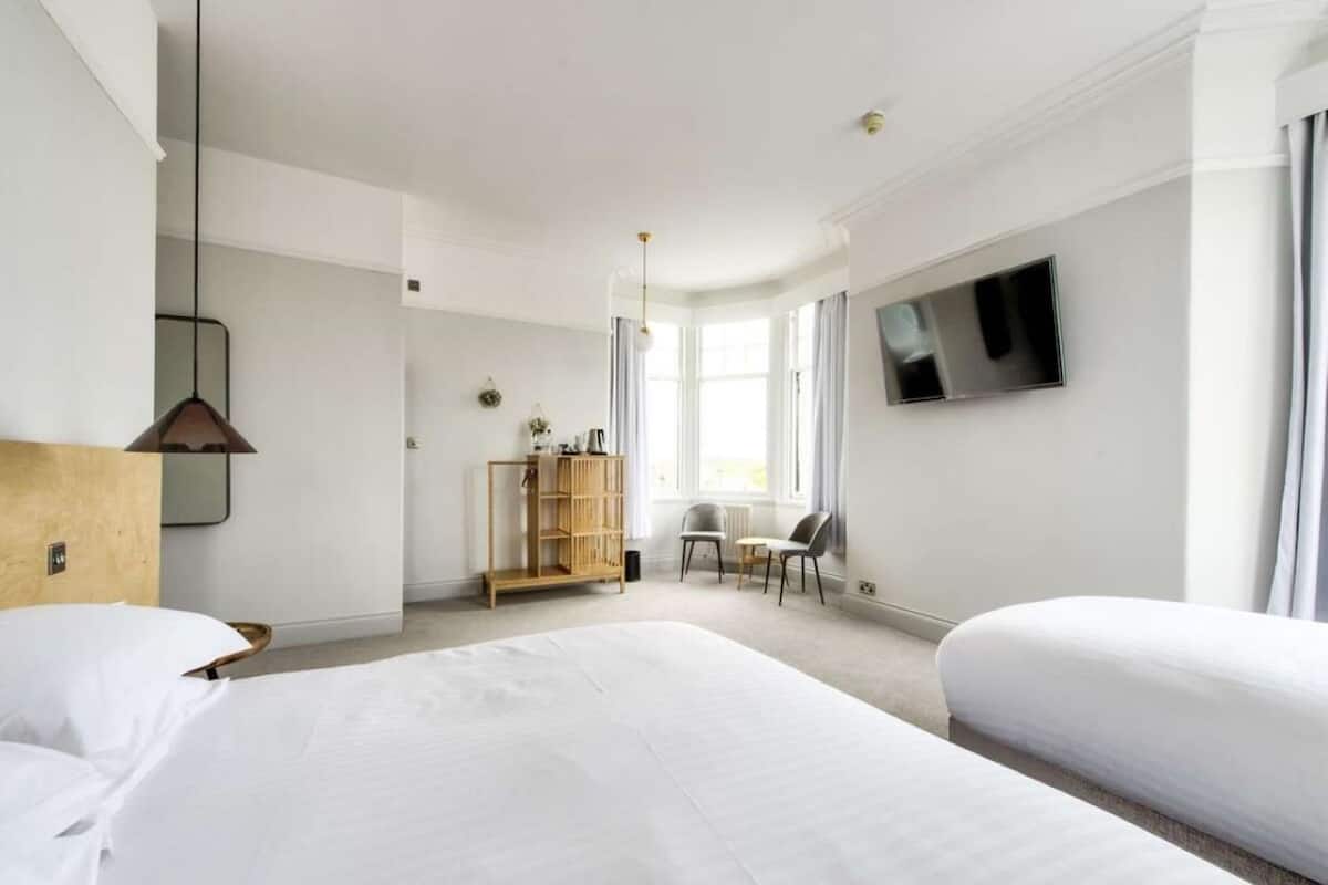 A hotel room with two beds, a TV mounted on the wall, a wooden wardrobe, and a seating area with a chair and a small table.