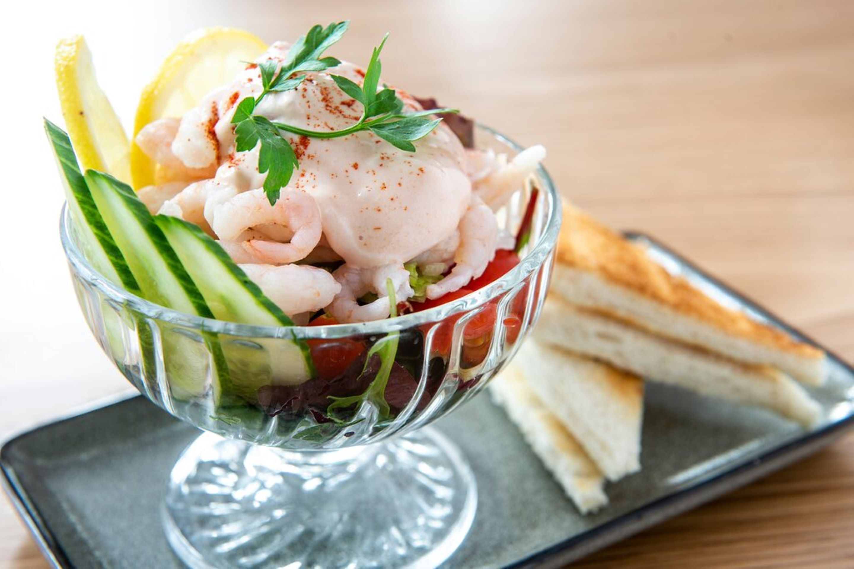 Een vissalade in een glazen schaal met citroenpartjes, komkommer en peterselie, vergezeld van geroosterd brood op een bord.