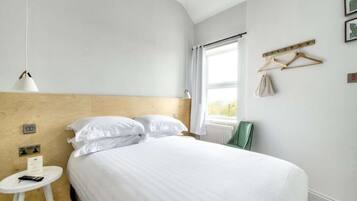 A hotel room with a bed, a small table with a lamp, a window with curtains, and a coat rack with a coat and bag.