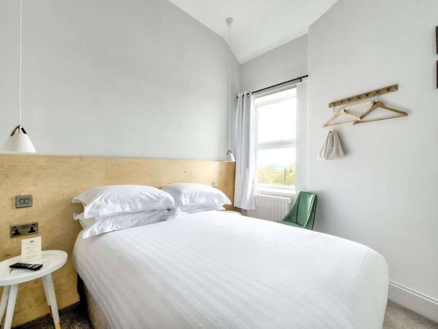 A hotel room with a bed, a small table with a lamp, a window with curtains, and a coat rack with a coat and bag.