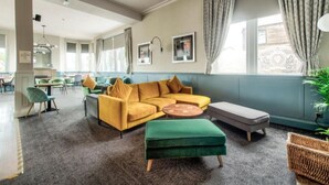 A modern living room with a yellow sofa, green ottoman, and a round coffee table. There are multiple chairs and a dining area in the background. - The Castle Hotel (Berwick-upon-Tweed)