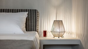 A neatly made bed with white pillows and a striped headboard, a bedside table with a glass of red drink, and a geometric lamp. - Golden Sand Villas (Naxos)