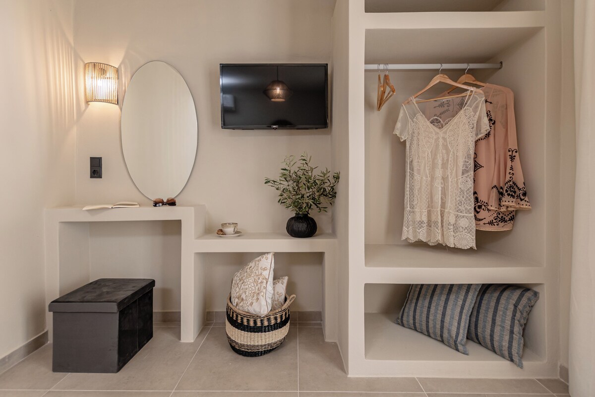 Habitación moderna con televisión de pantalla plana, un espejo grande, una mesita con una taza y un libro, una planta en maceta y un perchero con vestidos.