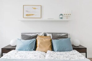 A neatly made bed with a floral-patterned bedspread, a framed artwork above the bed, and a wall-mounted shelf with a candle holder.