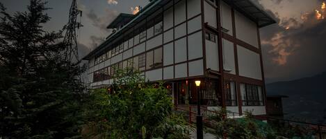 A traditional multi-story building with a prominent balcony, surrounded by lush greenery and a mountainous backdrop.