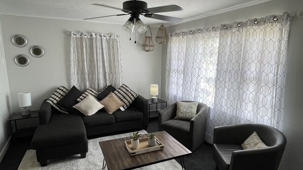 Un salon moderne avec un canapé noir, une table basse et un fauteuil gris. On y trouve un ventilateur de plafond, une lampe et une fenêtre avec des rideaux à motifs.