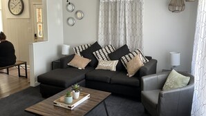 Un salon moderne avec un canapé gris foncé, une table basse en bois et un fauteuil gris. On y trouve également un ventilateur de plafond, une horloge murale et une fenêtre agrémentée de rideaux à motifs.