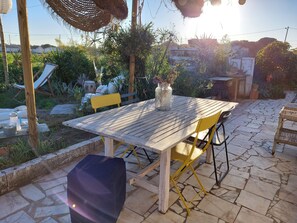 An outdoor patio with a wooden table and yellow chairs, surrounded by greenery and a stone floor.