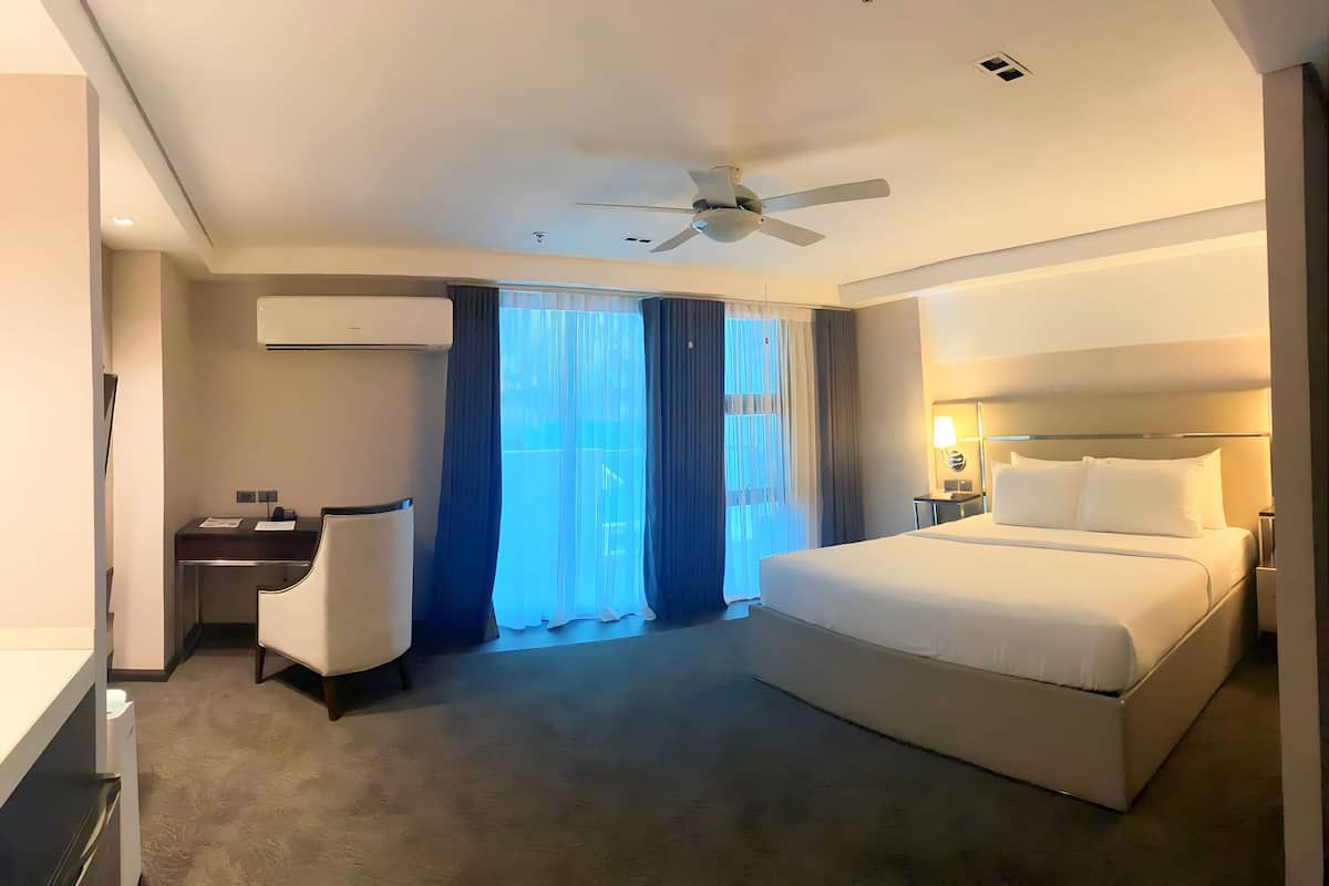 Habitación de hotel con una cama grande, un escritorio con silla, un ventilador de techo y una ventana con cortinas.
