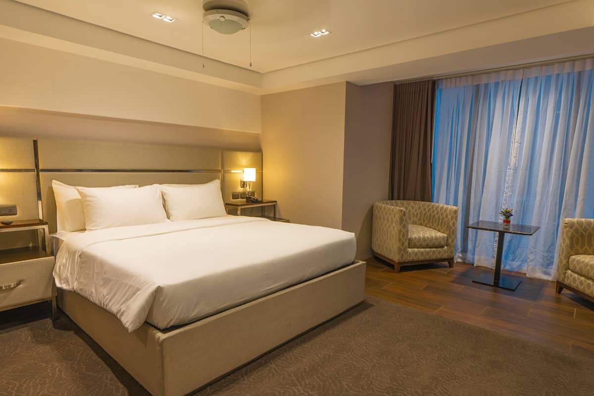 Habitación de hotel con una cama grande, dos mesitas de noche, una silla, una pequeña mesa y vistas al exterior a través de cortinas.