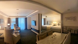 A modern hotel room with a flat-screen TV, a dining area with a table and chairs, a separate bedroom with a bed and bedside lamps, and a large window with curtains. - The Plaza Lodge Baguio (Baguio)