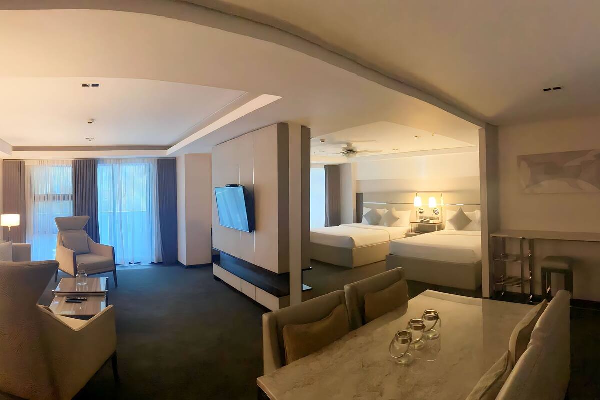 Una habitación de hotel moderna con televisor de pantalla plana, zona de comedor con mesa y sillas, dormitorio independiente con cama y lámparas de noche, y un ventanal con cortinas.