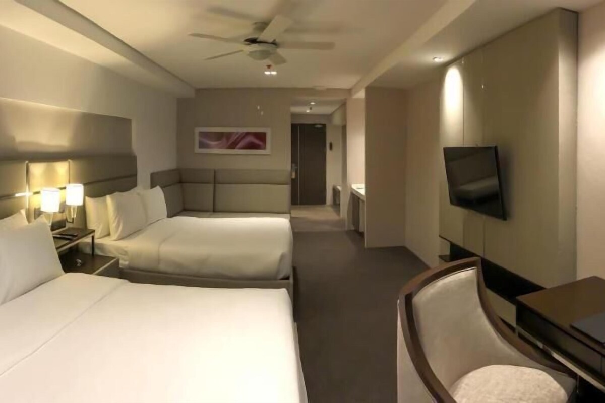 Habitación de hotel con dos camas, televisión, ventilador de techo y un pasillo que conduce a un baño.