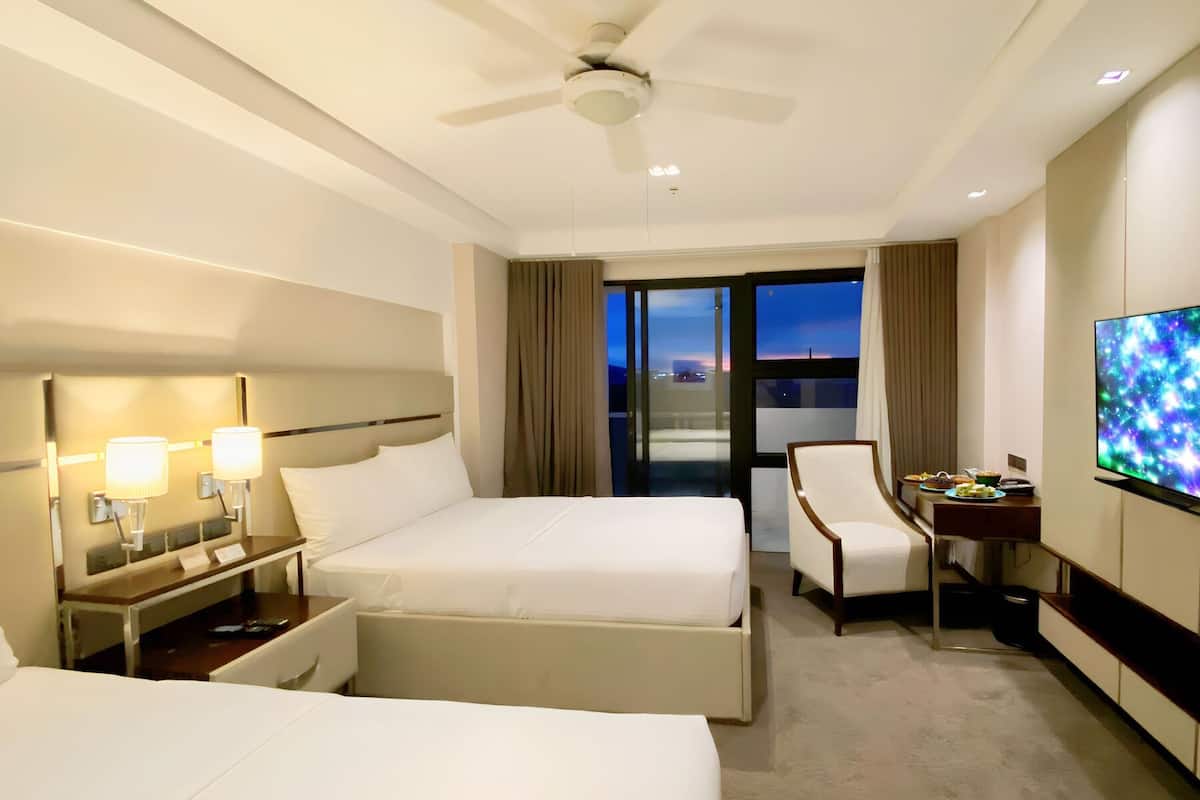 Habitación de hotel con cama, escritorio, silla, televisor y vista a la ciudad al atardecer.